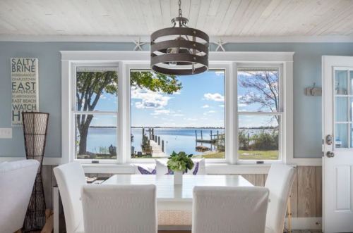 Bright dining area with a table, overlooking a serene lake through large windows, framed by trees and a dock.