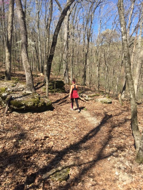 A person in a red outfit walks along a trail in a wooded area with trees and rocky terrain.