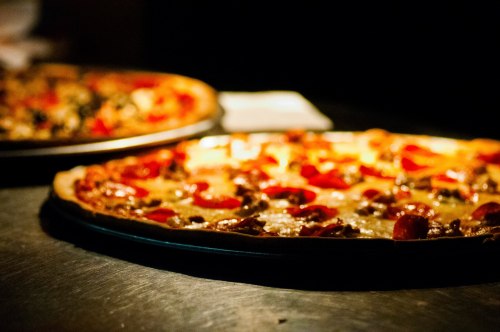 Two pizzas with pepperoni and toppings, placed on a dark surface, with one slightly in the foreground.