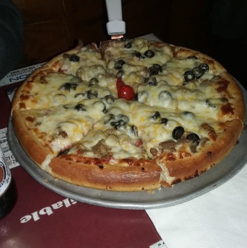 A round pizza topped with melted cheese, olives, sausage, and a cherry tomato, served on a metal tray.