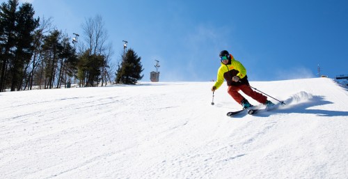 A skier in a bright yellow jacket glides down a snowy slope under a clear blue sky. Trees line the background.