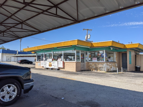 A small fast-food restaurant with a yellow awning, featuring large windows and a parking area. Clear blue sky above.