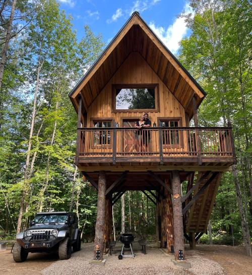 A wooden treehouse with a person on the balcony, surrounded by trees, and a black Jeep parked nearby.
