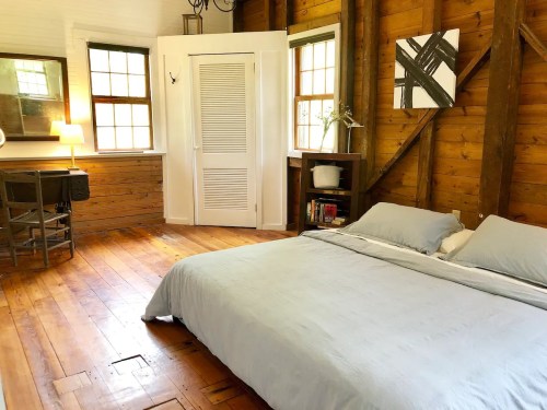 Cozy bedroom with wooden walls, a bed with light blue bedding, a desk, and large windows letting in natural light.