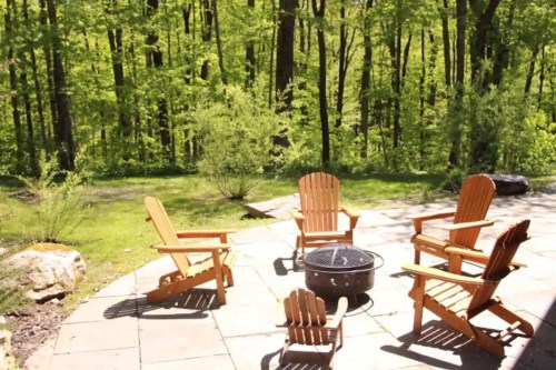 A serene outdoor patio with wooden chairs surrounding a fire pit, set against a lush green forest backdrop.