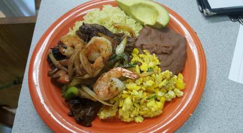 A colorful plate featuring grilled shrimp, beef, scrambled eggs, rice, refried beans, and avocado slices.