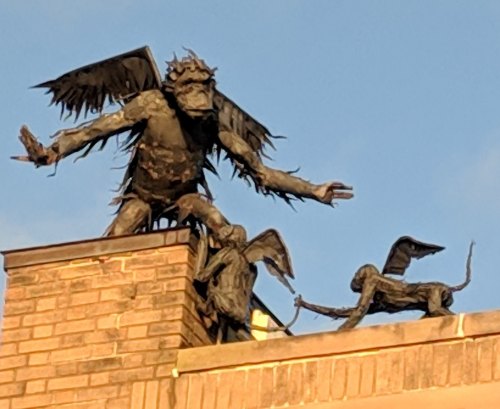 A bronze sculpture of a winged creature and two smaller figures on a brick rooftop against a blue sky.
