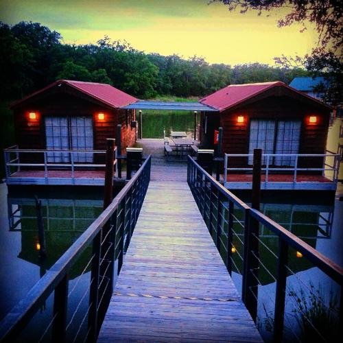 A wooden dock leads to two cozy cabins by a calm lake, surrounded by trees at dusk. Warm lights glow from the cabins.