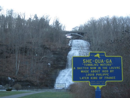A waterfall cascades down a rocky cliff, with a historical sign in the foreground detailing its name and history.