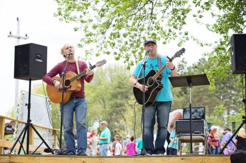 Two musicians perform on stage with acoustic guitars, surrounded by an audience enjoying the outdoor event.