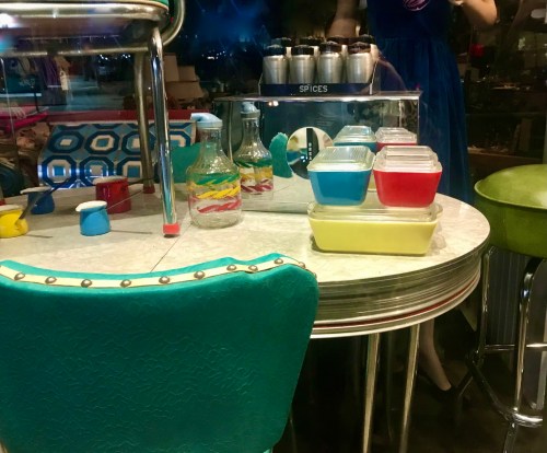 A retro diner table with colorful containers, glass bottles, and a green chair, reflecting a nostalgic vibe.