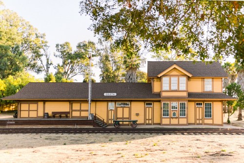 A historic yellow train station surrounded by trees, with a platform and vintage details, set in a sunny landscape.