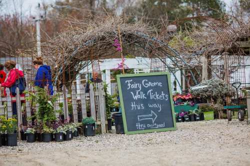 A sign directing visitors to Fairy Gnome Walk tickets, surrounded by plants and people at a garden event.