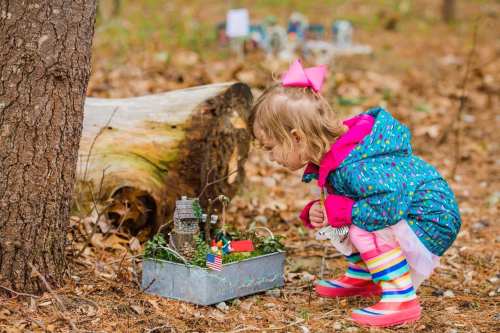 A young girl in a colorful coat and bow examines a small, whimsical outdoor display near a tree stump.