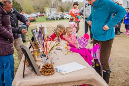 A group of children and adults engage in a craft activity at an outdoor event, with tables displaying art supplies.