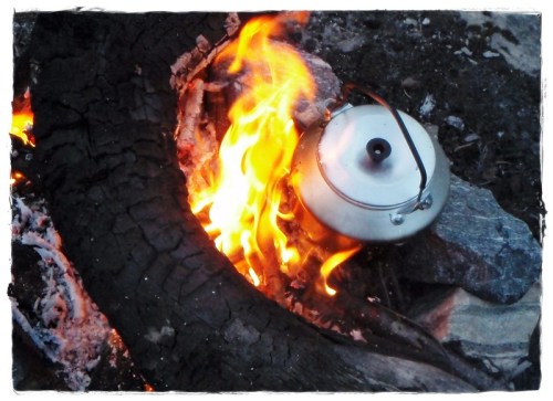 A kettle sits on a campfire, surrounded by flames and glowing embers, with a charred log nearby.