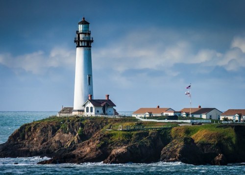 A tall lighthouse stands on a rocky shore, surrounded by ocean waves and a cloudy sky, with nearby buildings and a flag.