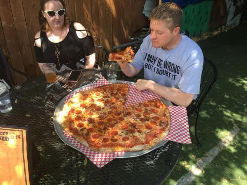 A man eats a large pepperoni pizza at an outdoor table, while a woman sits nearby with a drink.