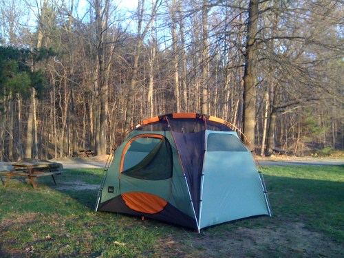 A tent set up in a wooded area, surrounded by trees and grass, with a picnic table visible in the background.