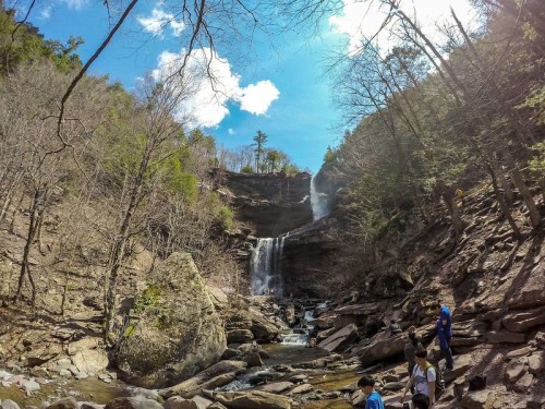 A scenic waterfall surrounded by rocky terrain and trees, with hikers exploring the area under a blue sky.
