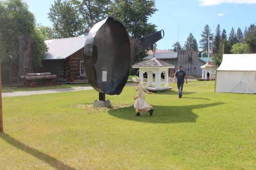 A large metal frying pan sculpture in a grassy area, with a child running and an adult walking nearby.