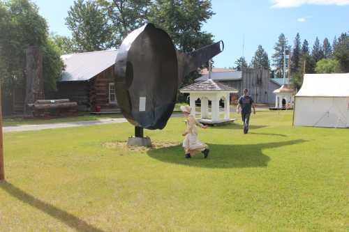 A large metal frying pan sculpture in a grassy area, with a child running and an adult walking nearby.