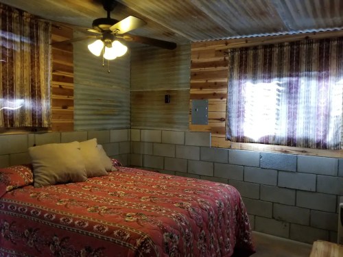 Cozy bedroom with a floral bedspread, ceiling fan, and rustic walls made of wood and cinder blocks. Natural light from windows.