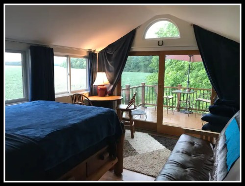 Cozy room with a bed, table, and large windows overlooking a green landscape and a deck with outdoor seating.