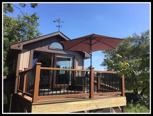 A cozy cabin with a deck, umbrella, and large windows, surrounded by trees and greenery.
