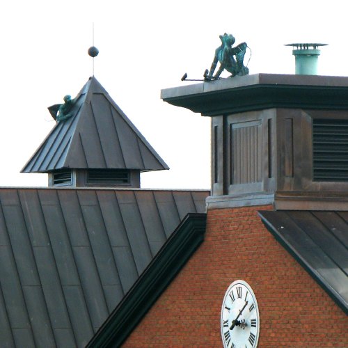 Two sculptures of workers on a building rooftop, one on a peak and the other near a clock, against a clear sky.