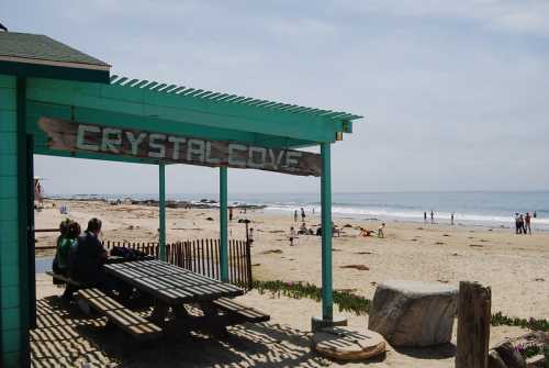 A beach scene at Crystal Cove with a wooden sign, picnic table, and people enjoying the sandy shore and ocean.