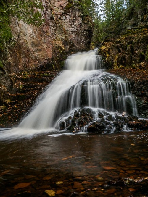 A serene waterfall cascades over rocky terrain, surrounded by lush greenery and autumn leaves.