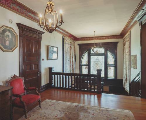 A vintage hallway with dark wood accents, a chandelier, and ornate stained glass windows, featuring a red armchair and rug.