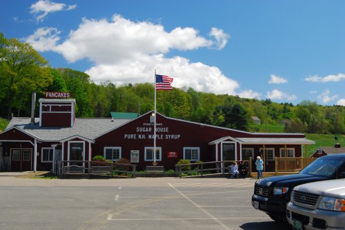 A red sugar house with a sign for pancakes and maple syrup, surrounded by green trees and a blue sky with clouds.