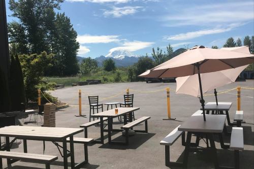 Outdoor seating area with tables and umbrellas, overlooking mountains and trees under a clear blue sky.