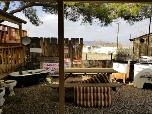 A rustic outdoor space with a wooden table, various signs, and eclectic furniture, surrounded by a wooden fence.