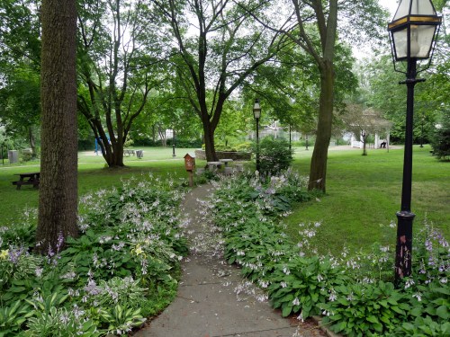 A serene park pathway lined with flowers, trees, and lampposts, leading to a grassy area with benches and a gazebo.