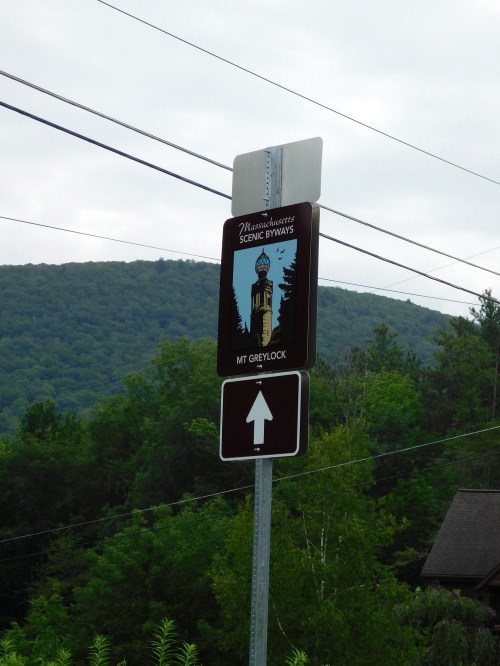 Sign for Massachusetts Scenic Byways directing to Mt. Greylock, with a backdrop of green mountains and cloudy sky.