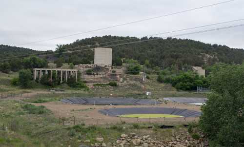 Abandoned industrial site with concrete structures, surrounded by greenery and a large circular area covered with black material.