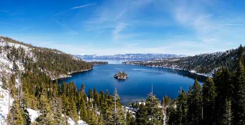 A panoramic view of a serene lake surrounded by snow-covered mountains and lush evergreen trees under a clear blue sky.