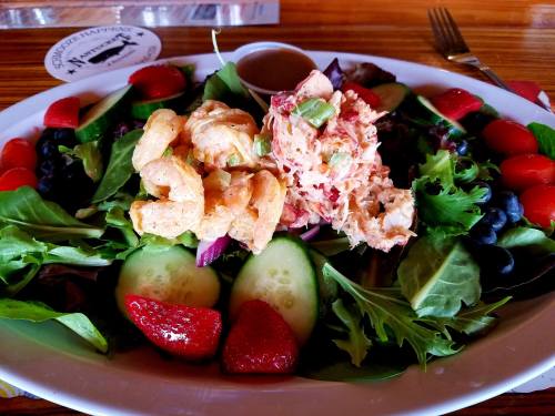 A colorful salad with shrimp, crab meat, cucumbers, strawberries, blueberries, and a dressing on the side.