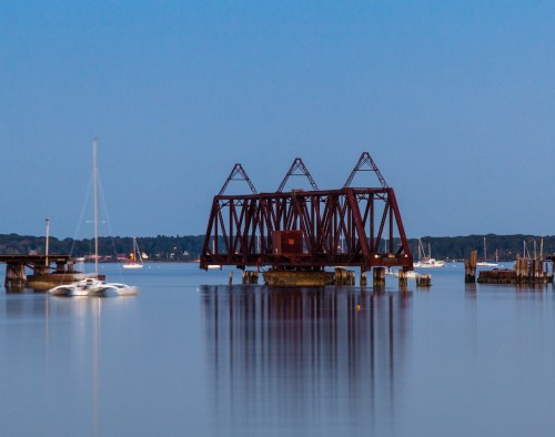 A rusted railway bridge stands over calm water, with boats and a distant shoreline under a twilight sky.