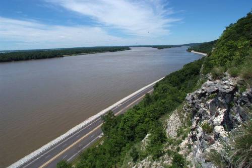A scenic view of a river winding through lush greenery, with a road along the riverbank under a clear blue sky.