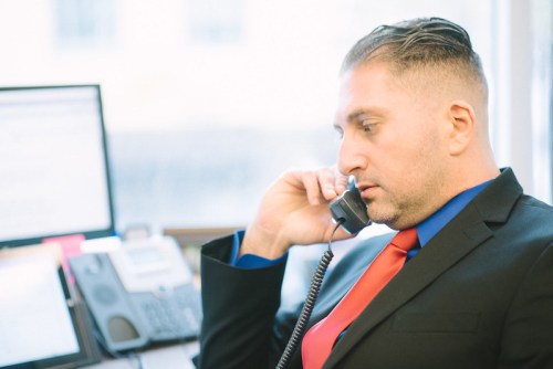 A man in a suit talks on the phone at an office desk with computers and office equipment in the background.