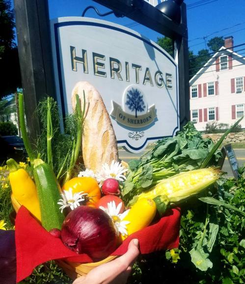 A hand holds a colorful basket of fresh vegetables and flowers in front of a sign for Heritage of Sherborn.