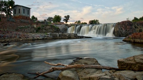 A serene waterfall cascades over rocks, with a historic building and trees in the background under a colorful sky.