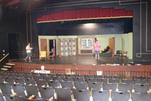 A theater stage with two people rehearsing, set design featuring a window and furniture, and empty audience seats.