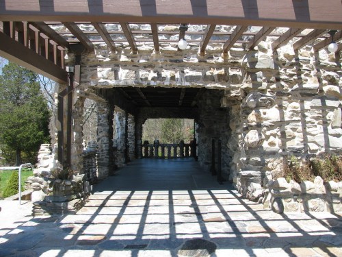 A stone structure with a wooden pergola, casting shadows on the ground, surrounded by greenery.