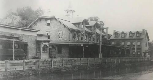 Black and white photo of a historic train station with a fence and nearby buildings.