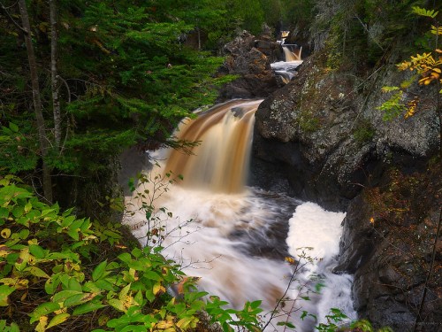 A serene waterfall cascades over rocky terrain, surrounded by lush green trees and autumn foliage.
